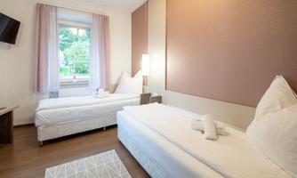 A bright hotel room with two single beds and a modern design. The room has a window with a view to the outside and a simple decor.
