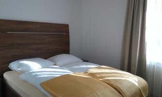 A bright bedroom with a double bed and white bed linens. Curtains and windows let natural light in.