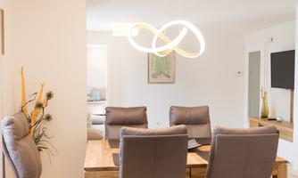 A modern dining room with a wooden chair set and elegant lighting. In the background, warm colors and decorative details can be seen.