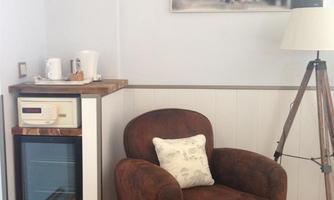 A cozy armchair is in a corner with a small table and a refrigerator. A picture hangs on the wall, and a lamp stands next to it.