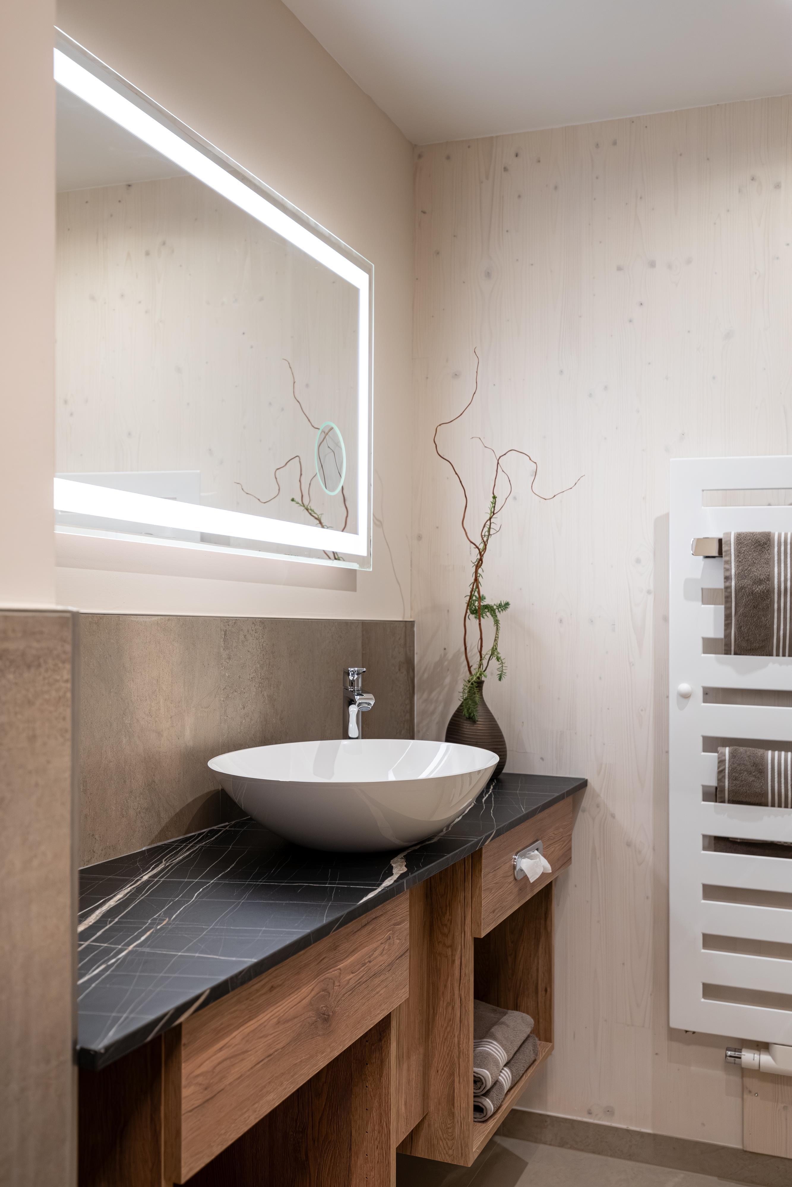 A modern bathroom with an elegant sink and illuminated mirror. The walls are in light tones and there is a subtle plant decoration.