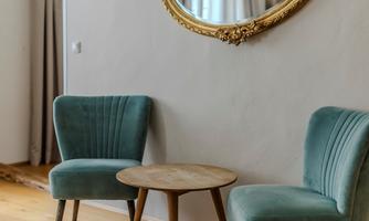 A cozy room with two turquoise chairs and a round table. An elegant, golden mirror hangs on the wall.