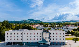 A modern hotel building in a green environment with gentle hills in the background. The sky is clear and blue, perfect for a stay in nature.