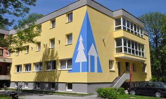 A light yellow building with large windows and a striking blue mural of trees. Surrounded by green trees and a clear blue sky.