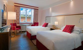 A modern hotel room with two single beds and a desk. The room has large windows overlooking the outside and is designed in bright colors.