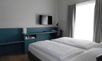 A modern room with a double bed and a flat-screen TV. The large windows provide plenty of daylight.
