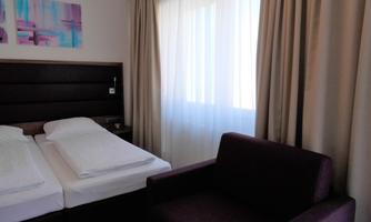 A bright and modern bedroom with two beds and a comfortable armchair. Large windows let in plenty of light.