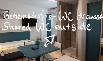 A modern room with a table, two chairs, and a single bed. The bathroom is separated by a glass wall.