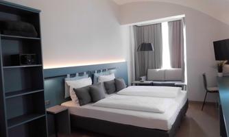 A modern bedroom with a double bed and gray pillows. The room is bright and minimally furnished, with a cozy seating area and large windows.
