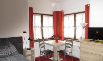 A cozy room with a bed, a small table, and chairs. Bright windows with red curtains create a friendly atmosphere.