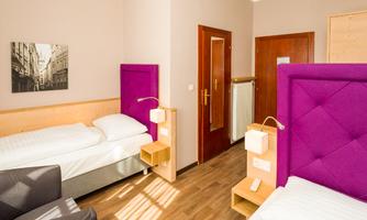 A modern hotel room with two single beds and stylish purple headboards. The decor is simple and cozy, with warm wood and neutral tones.