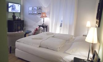 A cozy bedroom with a large bed and white bedcovers. In the room, there is a television, a floor lamp, and some pictures on the wall.