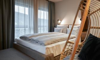 A modern bedroom with a large bed and bright curtains. A cozy seating area made of rattan complements the stylish atmosphere.