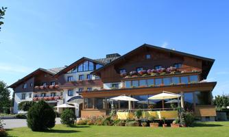 A beautiful hotel building with a wooden facade and balcony. The garden is well-kept, and a bright blue sky frames the scene.