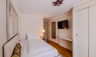 A modern bedroom with a single bed and minimalist furnishings. A television is hanging on the wall, and there is a cozy bedside lamp.