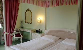 An elegant bedroom with a double bed and a decorative canopy. A mirror hangs on the wall, and a desk with a chair complements the furnishings.
