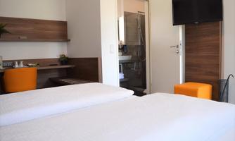 A modern hotel room with two beds and a television. In the background, a bathroom and a small table with a chair can be seen.