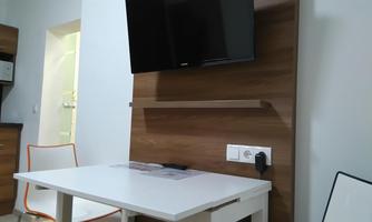A modern room with a white table and a television on the wall. In the background, wooden shelves and an example of a small kitchen corner are visible.
