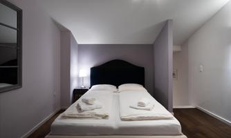 A modern bedroom with a large bed and fresh towels. The walls are painted in a soft purple, and there is a nightstand with a lamp.