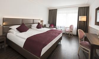 An elegant hotel room with two beds and a modern decor. Large windows provide plenty of daylight and a pleasant atmosphere.