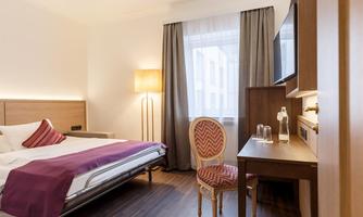 A modern hotel room with a double bed and an elegant desk. The large window front provides plenty of daylight.