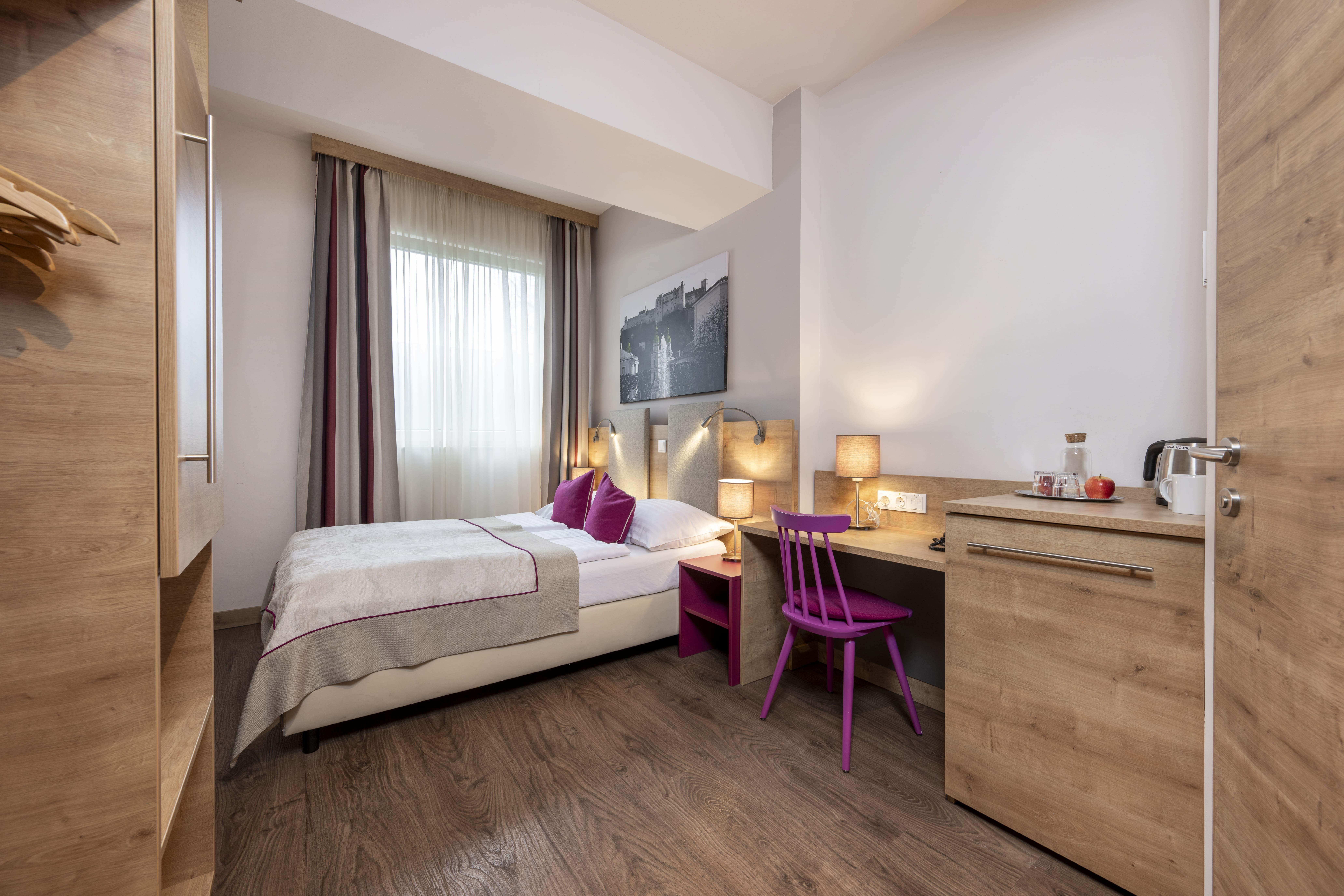 A modern hotel room with a single bed, desk, and a pink chair. Large windows provide ample daylight and a cozy atmosphere.