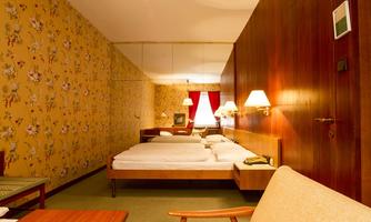 A cozy hotel room with two twin beds and an elegant wooden wall. The walls are decorated with a blooming wallpaper pattern, and there is a comfortable armchair as well as a table lamp.