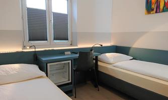 A modern guest room with two single beds and a table. There is a window with a view of the outside world and a small refrigerator.