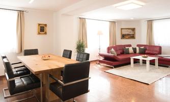 A modern living room with a red sofa and a white table. The dining area has a large wooden table with black chairs.