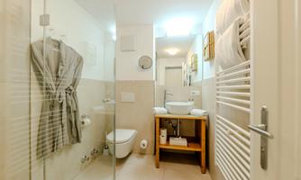 A modern bathroom with a glass shower and a sink. A bathrobe hangs on the wall and a wooden shelf with cosmetic products is ready.