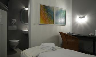 A simple room with a bed, a desk, and two paintings on the wall. In the background, a bathroom with a toilet is visible.