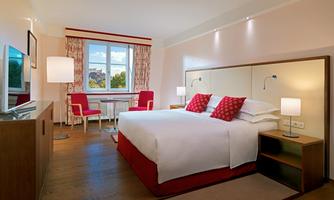 A modern hotel room with a large bed and red pillows. There is a desk and a window with a view outside.