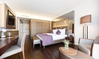 A modern hotel room with a large bed, elegant furnishings, and hardwood flooring. The room features a dining area, a coffee table, and a charming flower arrangement.