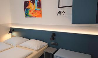 A modern bedroom with two beds and a stylish wall design.  
A colorful artwork hangs on the wall, and there is subtle lighting.