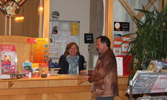 A reception area with a friendly staff member and a visitor. In the background, informational materials and plants are visible.