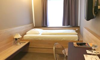 A simple bedroom with a single bed and a desk. The room has large windows with curtains and is decorated in neutral colors.