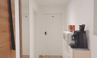 A bright hallway with a white wall and a wooden shelf. On the right side, there is a coffee machine and a refrigerator.