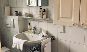 A modern bathroom with white tiles and a sink. Care products are on the sink and a towel is draped.