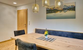 A modern dining area with a wooden table and comfortable chairs. Stylish lamps hang over the table, and a painting of a landscape adorns the wall.