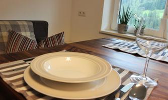 A laid table with a white plate, cutlery, and a glass. In the background, a cozy seating area and a windowsill with plants can be seen.