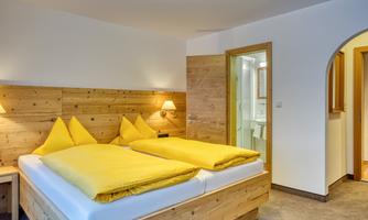 A cozy bedroom with two single beds and yellow pillows. In the background, wooden details and an adjacent bathroom are visible.
