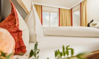 A cozy bedroom with a large bed and decorative pillows. The windows allow plenty of daylight in, and the curtains give the room a warm atmosphere.