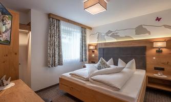 A modern bedroom with wooden furniture and a large window. The bedding is inviting, and the walls are decorated with subtle patterns.