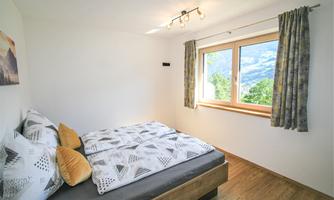 A bright bedroom with a double bed and a view of nature. The walls are white, and large windows let in plenty of light.