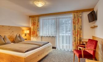 A cozy room with a double bed and warm wooden furniture. The room has a window with curtains and a television on the wall.