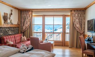 A cozy hotel room with wooden details and a view of the mountains.  
Large windows let in a lot of daylight and showcase the snow-covered landscape.
