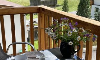 A cozy balcony with a table on which coffee, dishes, and a vase of flowers are placed. In the background, green meadows and trees can be seen.