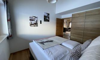 A cozy bedroom with a double bed and wooden shelves. Pictures hang on the wall, and the window lets in plenty of daylight.