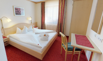 A cozy hotel room with a large bed and two pillows. There is a desk and a chair as well as a bright window front.
