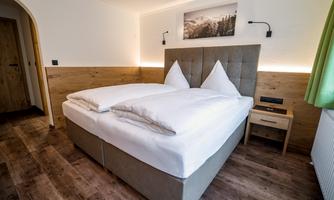 A modern bedroom with a double bed and white pillows. The walls are bright, and there is a green curtain as well as a nightstand next to the bed.